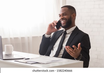 Excited Young Black Businessman Talking On Mobile Phone With Friends In Modern White Office, Planning Holidays Or Weekend, Copy Space