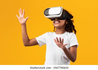 Excited young black girl in VR headset touching air during virtual reality experience at studio. Emotional kid exploring artificial computer world or playing video game, isolated on yellow background