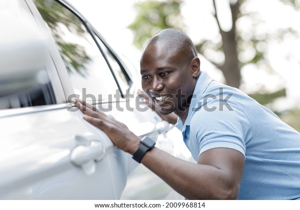 Excited young\
black man buying new car, standing on the street and checking\
automobile surface, closeup. Smiling african american guy cleaning\
his beautiful brand new white auto\
outdoors