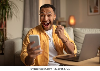 Excited Young Black Man Looking At Smartphone Screen, Gesturing YES, Feeling Happy About Online Win At Home. Millennial Guy Winning Lottery Or Casino Bet, Video Gaming On Cellphone