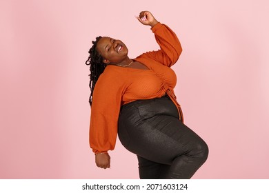 Excited young black plus size body positive woman with dreadlocks in orange top dances standing on light pink background in studio closeup - Shutterstock ID 2071603124