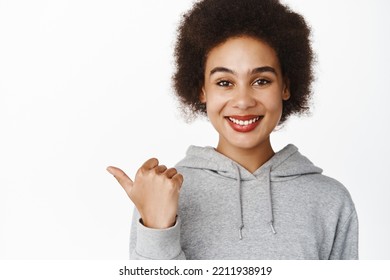 Excited Young Black Woman With Afro Hair, Wearing Grey Hoodie, Pointing Finger Left, Showing Advertisement, Promo Banner On Copy Space, Company Logo, White Background