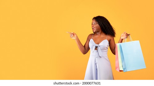 Excited young black woman with bright shopping bags pointing aside, offering big sale or unexpected promo on orange studio background, copy space. Seasonal discounts, offers, deals concept