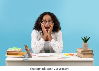 Excited Young Black Woman In Glasses Sitting At Desk With Study Materials, Achieving Success In Exams On Blue Studio Background. Millennial African American Woman Celebrating Victory, Copy Space