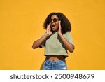 Excited young Black woman wearing sunglasses standing at yellow wall. Happy surprised African American gen z girl wow face feeling surprise looking away advertising summer shopping sale, travel offer.