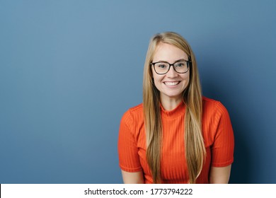 Excited Young Blond Woman Wearing Spectacles In A Colorful Orange Top Grinning Happily At The Camera Over A Blue Studio Background With Copy Space