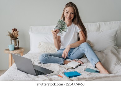 Excited young blonde girl in jeans in casual sitting on bed with laptop, notebook, phone, diary, holds dollar banknotes in fan shape, happily looks at camera, satisfied by salary. Young American busin - Shutterstock ID 2254774463