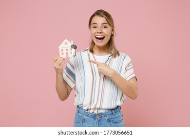 Excited young blonde woman girl in casual striped shirt posing isolated on pastel pink background studio. People lifestyle concept. Mock up copy space. Pointing index finger on house, bunch of keys