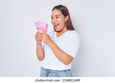 Excited young blonde woman girl Asian wearing casual white t-shirt looking at money rupiah banknotes in hand and rejoicing success isolated on white background. people lifestyle concept - Shutterstock ID 2248811489