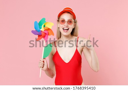Excited young blonde woman in red one-piece swimsuit cap eyeglasses isolated on pink background. People summer vacation rest lifestyle concept. Mock up copy space. Hold toy windmill, showing thumb up