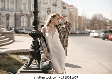 Excited Young Blonde Woman In Stylish Silk White Dress, Checkered Tweed Coat And Pearl Necklace Smiles And Poses Outside.