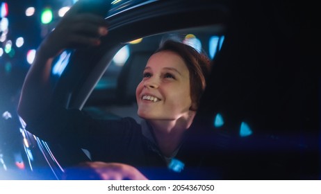 Excited Young Boy Is Looking Out Of The Window From Backseat Of A Car At Night. Kid Taking Photos And Videos On A Smartphone With Amazement Of How Beautiful Is The City Street With Working Neon Signs.