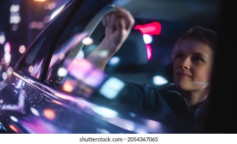 Excited Young Boy Is Sitting On Backseat Of A Car, Commuting Home At Night. Looking Out And Drawing On The Taxi Window While Riding The City Street With Working Neon Signs.