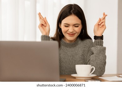 Excited young brunette businesswoman sitting in front of laptop with crossed fingers and closed eyes, have hope for good news, successful business meeting, drinking coffee at home office - Powered by Shutterstock