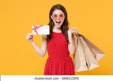 Excited Young Brunette Woman 20s In Red Summer Dress Eyeglasses Posing Isolated On Yellow Background Studio. People Lifestyle Concept. Hold Package Bag With Purchases After Shopping Gift Certificate