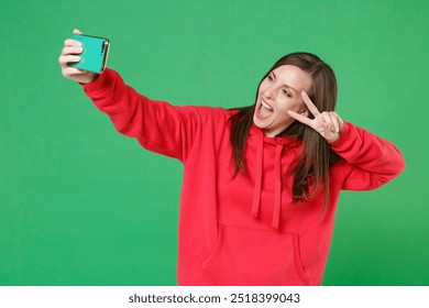 Excited young brunette woman 20s wearing bright red casual streetwear hoodie posing doing selfie shot on mobile phone showing victory sign isolated on green color wall background studio portrait - Powered by Shutterstock