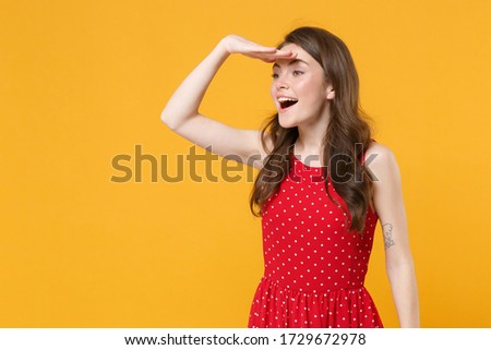Excited young brunette woman girl in red summer dress posing isolated on yellow background in studio. People lifestyle concept. Mock up copy space. Holding hand at forehead looking far away distance