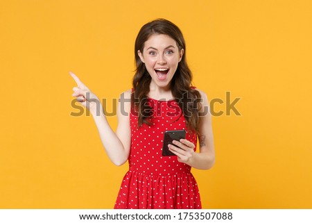 Excited young brunette woman girl in red summer dress posing isolated on yellow background in studio. People lifestyle concept. Mock up copy space. Using mobile phone, pointing index finger aside up