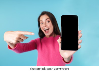 Excited Young Brunette Woman Standing Isolated Over Blue Background, Showing Blank Screen Mobile Phone, Pointing Finger