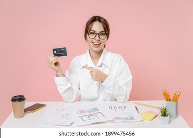 Excited Young Business Woman Employee In White Shirt Glasses Point Index Finger On Credit Bank Card Work In Office Sit At Desk Isolated On Pink Background Studio. Achievement Business Career Concept