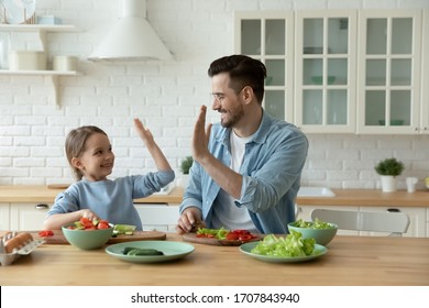 Excited Young Caucasian Father And Little Daughter Give High Five Engaged In Funny Activity Cooking Salad At Home Together, Happy Dad And Small Girl Child Have Fun Preparing Healthy Food In Kitchen