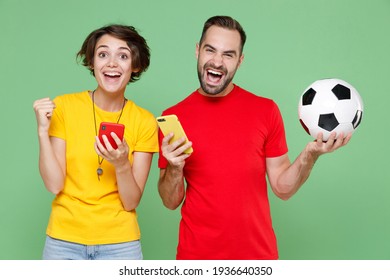 Excited Young Couple Friends Sport Family Woman Man Football Fans In T-shirts Cheer Up Support Favorite Team With Soccer Ball Using Mobile Phones Doing Winner Gesture Isolated On Green Background