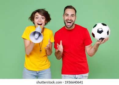Excited young couple friends sport family woman man football fans in t-shirts cheer up support favorite team with soccer ball scream in megaphone spreading hands isolated on green background studio