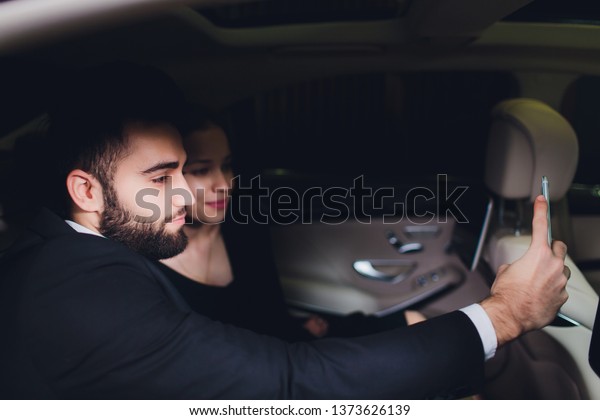 Excited young couple making a\
decision to purchase a new car sitting inside in a motor showroom\
checking it out for ownership and make selfie picture using\
smartphone.