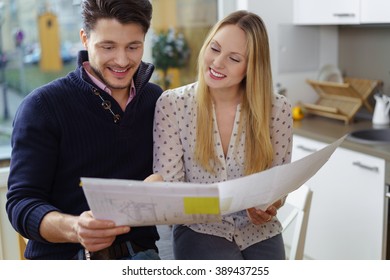 Excited Young Couple Making Plans For A New Home Standing In A Kitchen Holding Blueprint Designs For A Renovation Or New Build