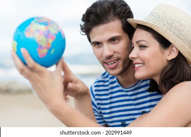 Excited Young Couple Planning Their Holiday Holding Up A Globe And Pointing To A Travel Destination With Focus To The Attractive Woman In A Trendy Straw Hat