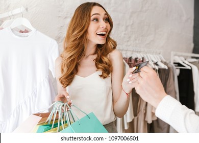 Excited Young Couple Shopping For Clothes Together At The Clothing Store, Woman Holding Lots Of Shopping Bags And Man Giving Credit Card