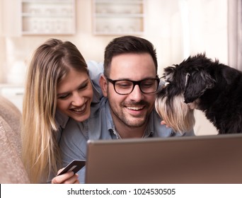 Excited Young Couple Shopping Online, Holding Credit Card And Looking At Laptop On Sofa At Home While Dog Making Their Company