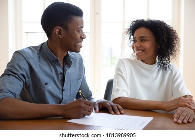Excited young couple sit at desk put signature on contract feel euphoric buying first house together, smiling African American husband and wife look at each other sign agreement at realtor office