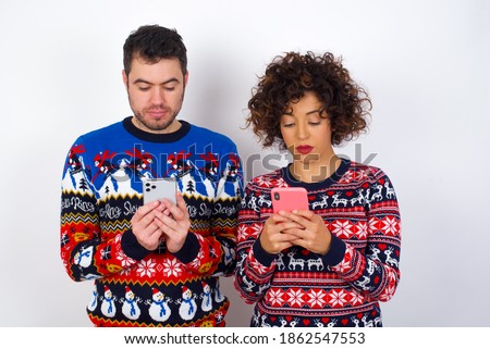 Excited Young couple wearing Christmas sweater standing against white background  winking and eye hold smart phone use read social network news