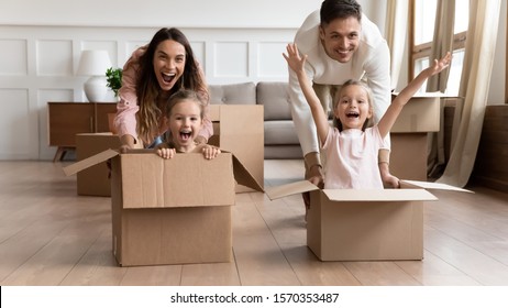 Excited Young Family Having Fun On Moving Day, Happy Parents Pushing Cardboard Boxes With Funny Cute Kids Daughters Riding In Containers Looking At Camera Laughing Celebrate Relocation Into New Home