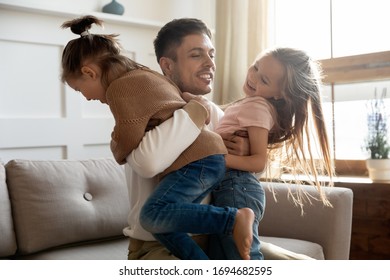 Excited young father holding two playful kids siblings, sitting on couch. Happy daddy having fun playing enjoying free weekend leisure time with energetic small children daughters in living room.
