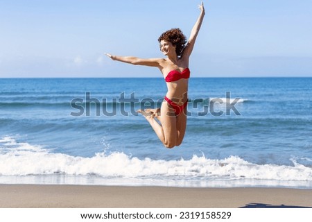 Excited young female in pink bikini stretching arms and jumping in air with folded legs up, while smiling and looking away in daylight against rippling sea and blue sky