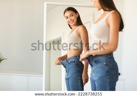 Excited young fit woman losing weight and wearing old too big jeans, woman feeling satisfied with results of her diet and slimming, posing near mirror, free space