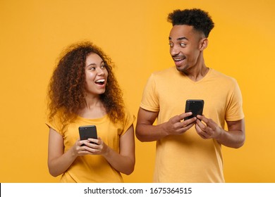 Excited Young Friends Couple African American Guy Girl In Casual Clothes Isolated On Yellow Orange Background. People Lifestyle Concept. Mock Up Copy Space. Using Mobile Phone, Looking At Each Other