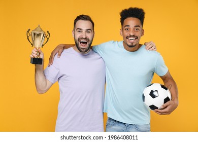 Excited Young Friends European African Men Football Fans In T-shirts Cheer Up Support Favorite Team With Soccer Ball Golden Cup Hug Isolated On Yellow Background Studio Portrait. Tattoo Translate Fun