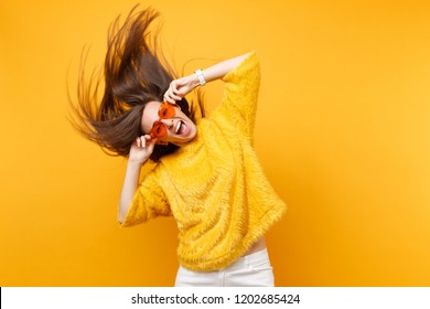 Excited Young Girl In Fur Sweater Holding Heart Orange Glasses Fooling Around In Studio Jumping With Fluttering Hair Isolated On Yellow Background. People Sincere Emotions Lifestyle. Advertising Area