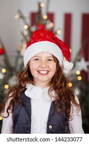 Excited Young Girl In A Red Santa Hat Standing In Front Of A Christmas Tree Decorated With Twinkling Lights In The Living Room