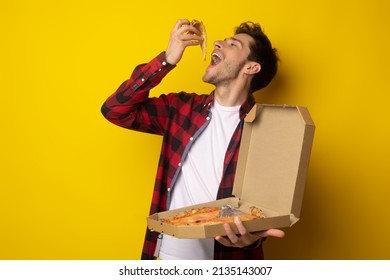 Excited Young Guy Enjoying Pizza Holding And Biting Tasty Slice Posing With Open Mouth Holding Carton Box On Yellow Orange Studio. Junk Food Lover Eating Italian Pizza. Unhealthy Nutrition Cheat Meal