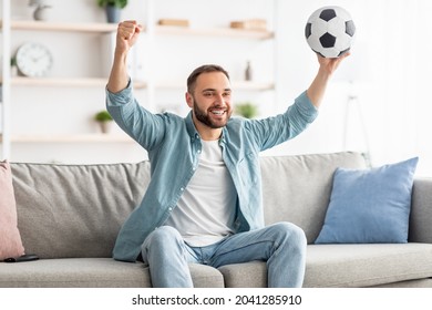 Excited Young Guy Watching Football On TV, Lifting Ball Above His Head, Celebrating Goal Or Victory On Couch At Home. Happy Millennial Man Supporting Favorite Soccer Team, Watching Championship