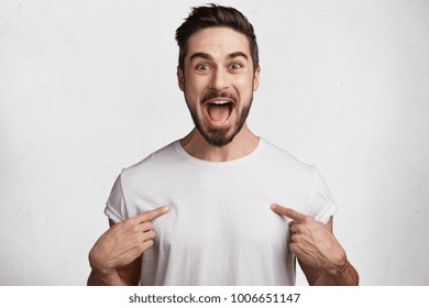 Excited young handsome male brunet with trendy hairdo, mustache and beard, happy to buy new clothes, indicates at blank space of t shirt for your advertising content, promotional text or logo.