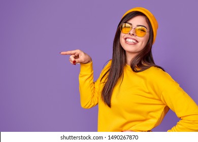 Excited young hipster in bright yellow outfit smiling and looking at camera while pointing at empty space on violet background - Shutterstock ID 1490267870