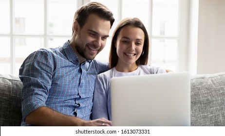 Excited Young Husband And Wife Sit Relax On Couch In Living Room Shopping On Internet Using Laptop At Home, Happy Millennial Couple Spouses Rest On Sofa Watching Funny Video On Computer Together