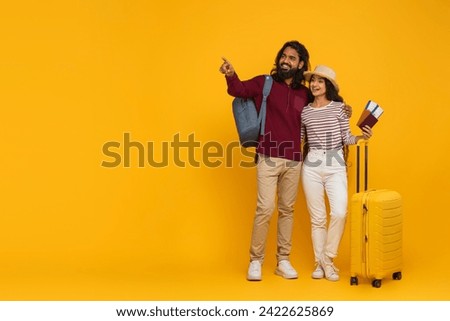 Excited young indian couple tourists going vacation, carrying backpack, suitcase, passport and flight tickets, pointing at blank copy space, showing travelling offer, yellow background