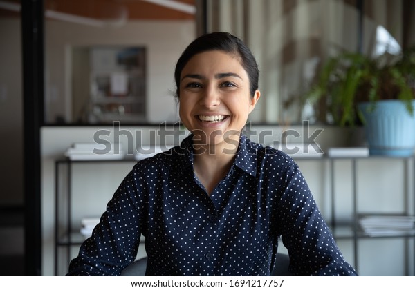 Excited young indian ethnicity woman looking at\
camera, holding funny conversation with colleagues online, webcam\
view. Head shot millennial hindu female worker laughing, having fun\
in office.