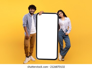 Excited young indian man and woman leaning on giant smartphone with mockup free space, promoting app or website, standing over yellow studio background - Shutterstock ID 2126870132